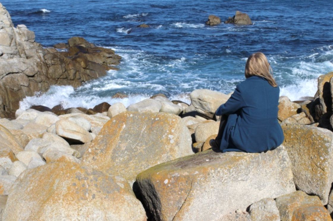 woman in blue coat with blond hair sits on large gray rock covered in yellow lichen watching blue water crash at into white spray at her feet