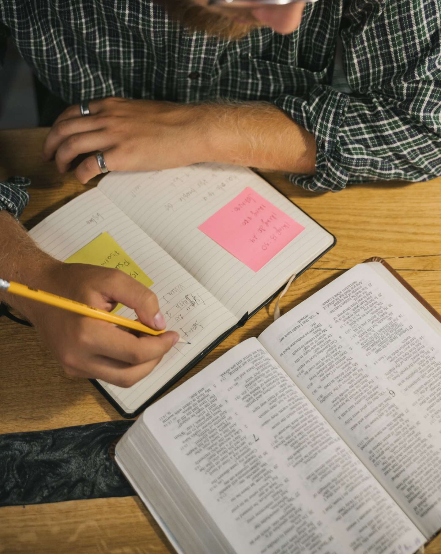 young adult male writes with pencil in notebook at wooden desk and refers to open Bible book