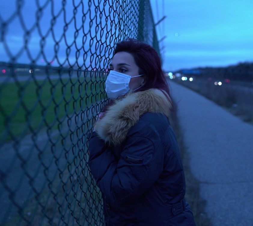 woman in face mask over her nose and mouth looking through a chain link fence