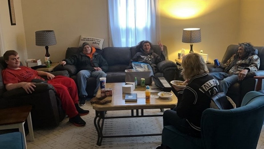 Five persons sit in overstuffed living room furniture around a coffee table.