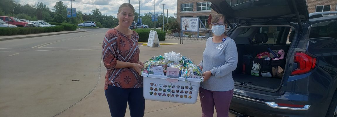 female charity volunteer and female hospital employee stand on either side of a plastic laundry basket full of edible gifts, ribbons and greeting cards of encouragement