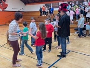 adults and children stand on school gym floor while other people stand in school bleachers