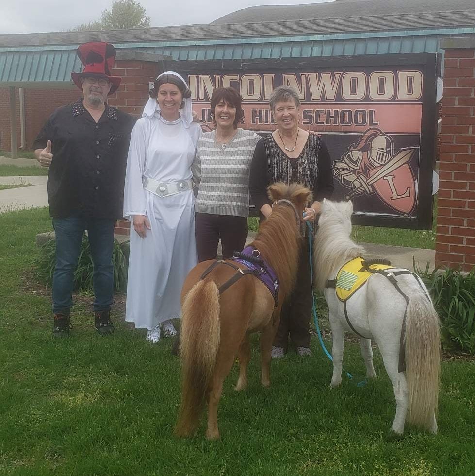man in tall red hat, woman in white dress, woman in striped sweater, woman in black shirt, brown mini horse in purple harness and white mini horse in yellow harness