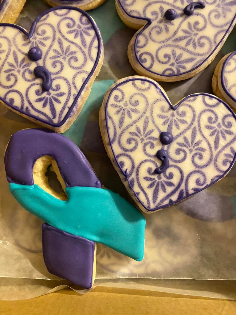 three cookies in cut out shapes of purple hearts with semi-colon icing and purple and teal cause ribbons.