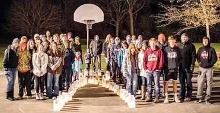 Forty people dressed in warm clothes stand outdoors on concrete behind white lunch bags lit from within with electric candles, all the bags arranged in the shape of a ribbon twist for a cause.
