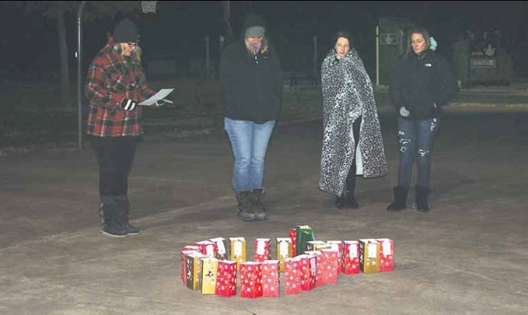 Four women dressed in warm clothes stand outdoors at nighttime on concrete behind red, gold, and green lunch bags lit from within with electric candles, all the bags arranged in the shape of a ribbon twist for a cause.