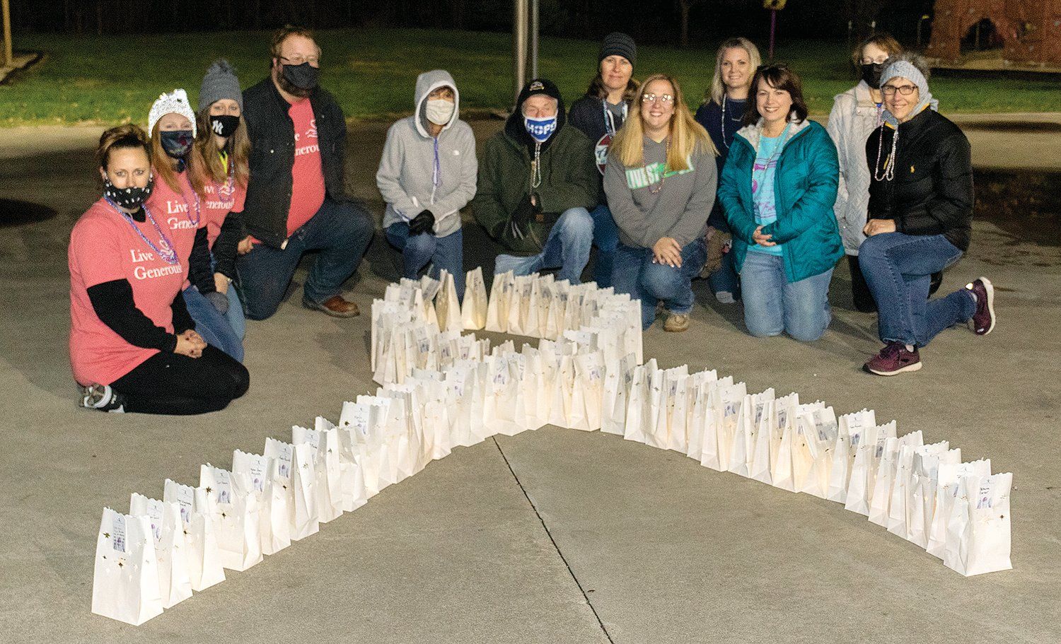 Twelve people dressed in warm clothes kneel outdoors behind white lunch bags lit from within with electric candles, all the bags arranged in the shape of a ribbon twist for a cause.