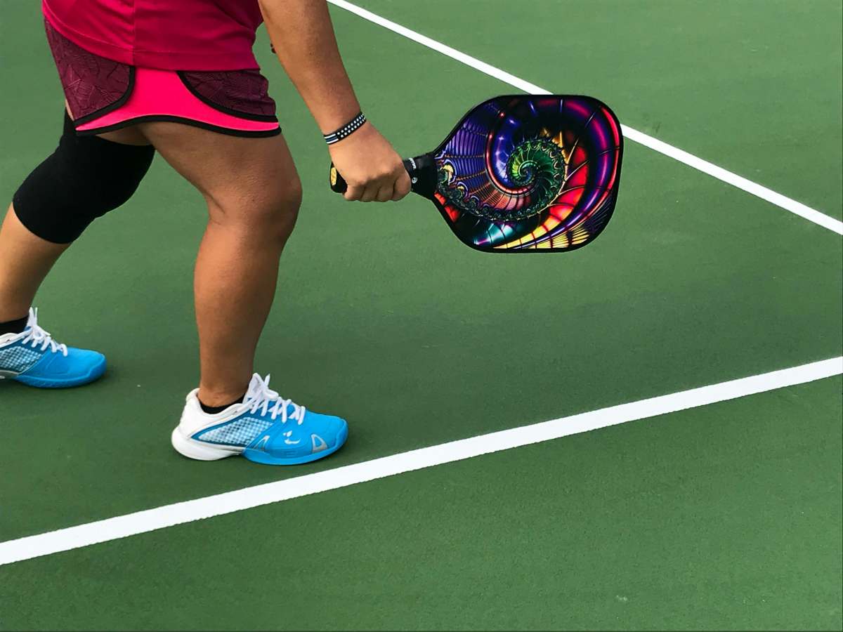 young person in shorts, blue tennis shoes and knee brace is on a green hard court ready to swing colorful pickleball racket.