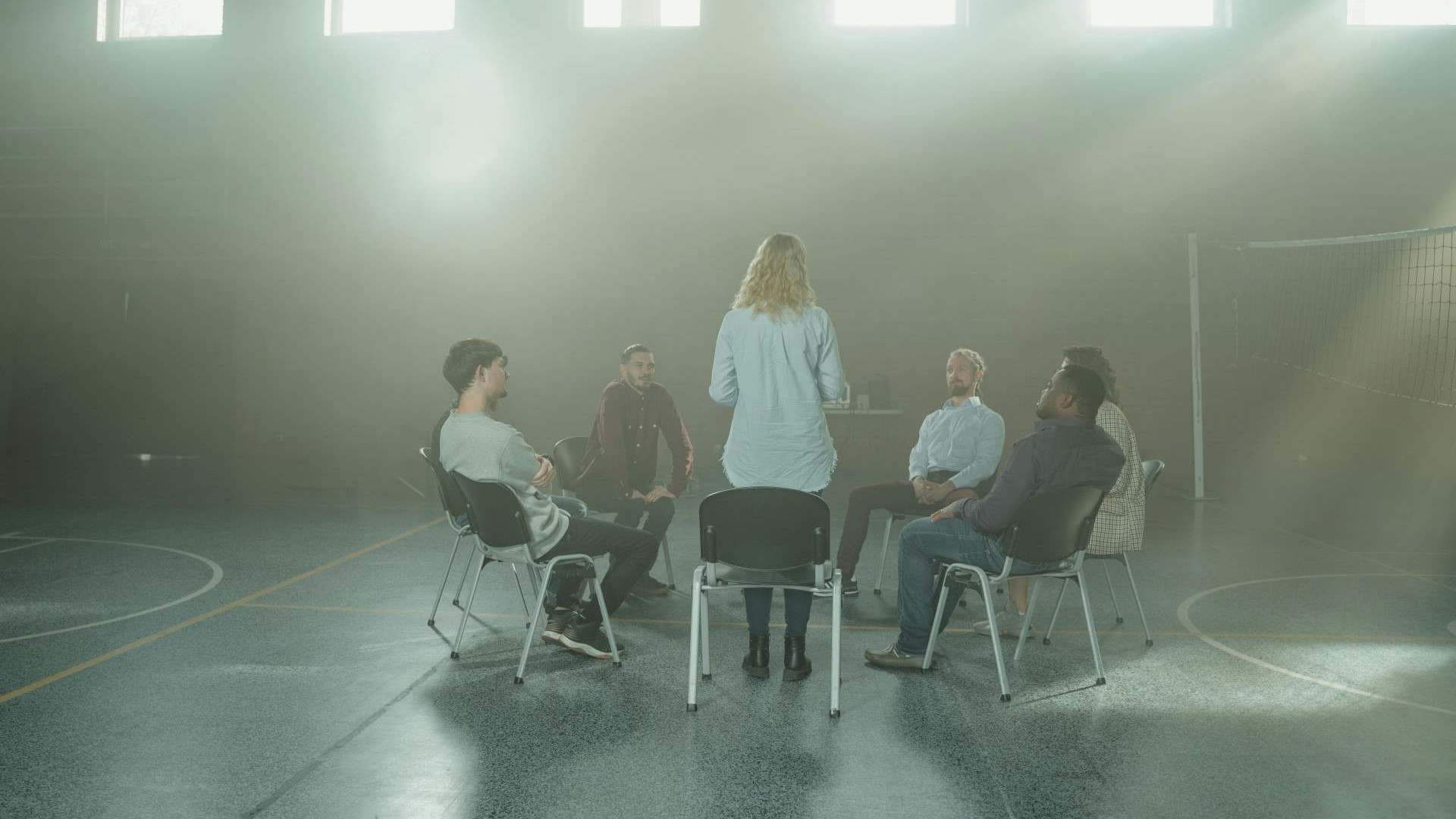 six persons sit in circle of simple chairs while one woman stands to share