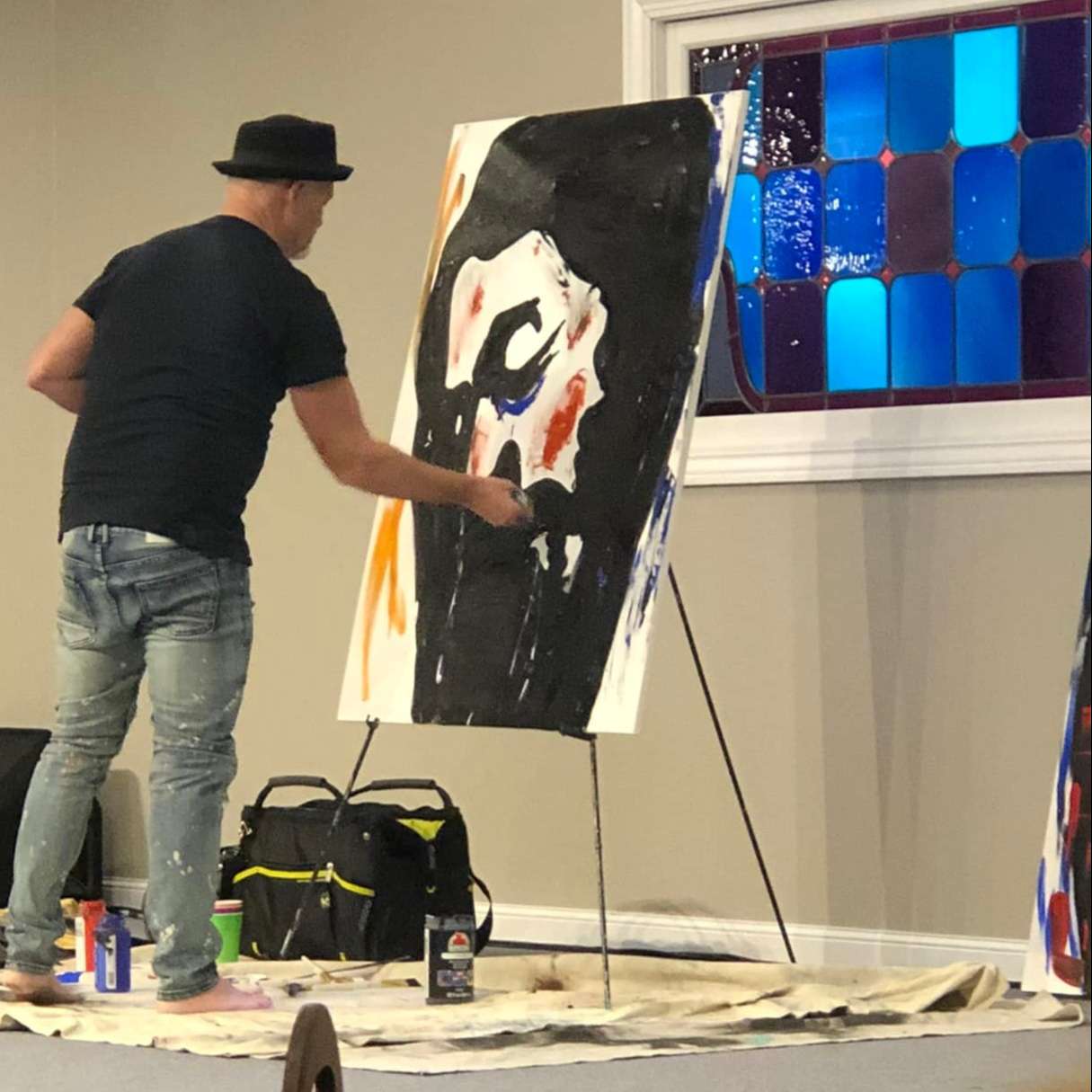 man in black derby hat, black tee shirt, and jeans, barefoot on dropcloth paints an image of Jesus Christ on a large canvas