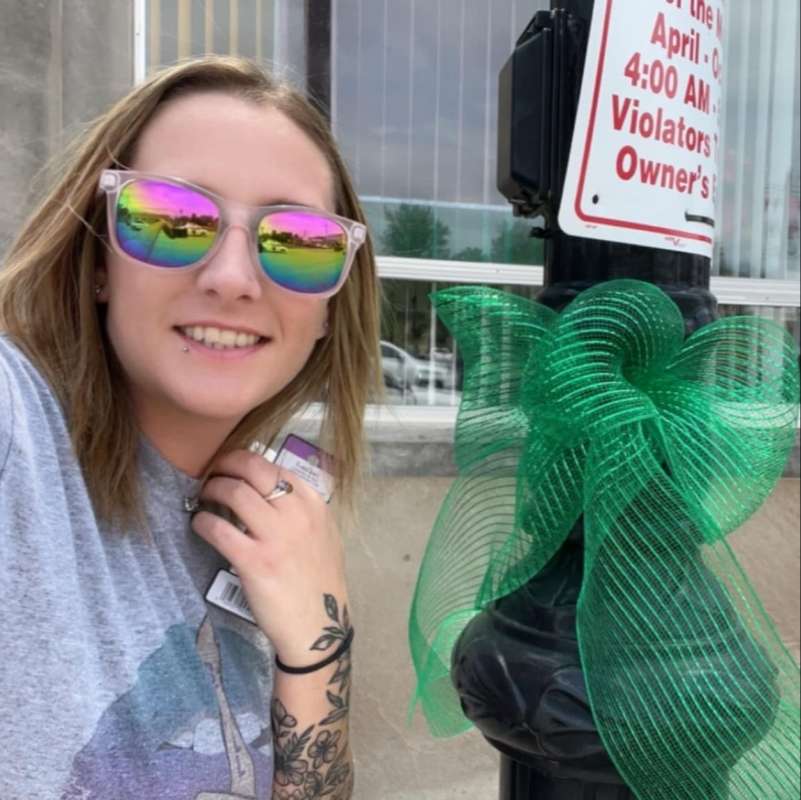 Woman in reflective sunglasses poses with a large green bow she's just tied around a city lamp post