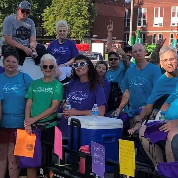 women and men of all ages wearing purple, blue, or green tee shirts, some stand on the street and some sit on a low trailer pulled by a pick up truck