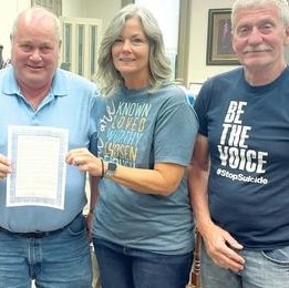 three middle aged people in blue shirts and blue demin jeans hold certificate declaring May as Mental Health Awareness Month in the county