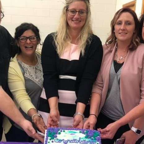 three adult ladies in cardigans and blazers smile and tilt up a sheet cake with purple icing