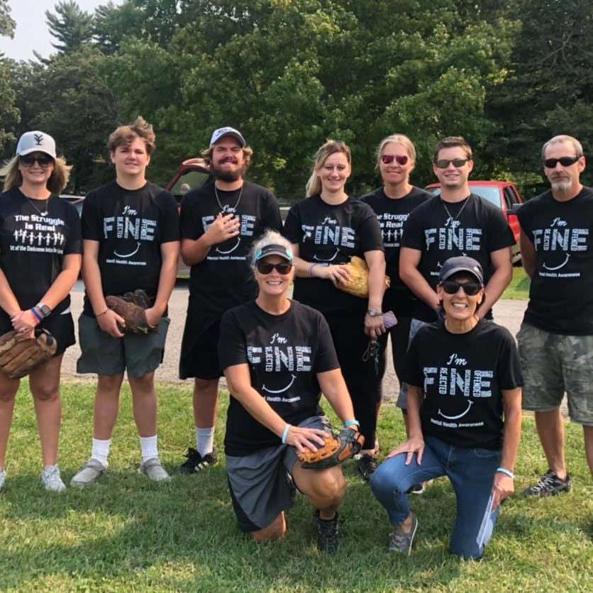 mixed gender group of people stand and kneel in the sun wearing softball gloves, sunglasses, and black team tee shirts that read “I'm Fine” 
